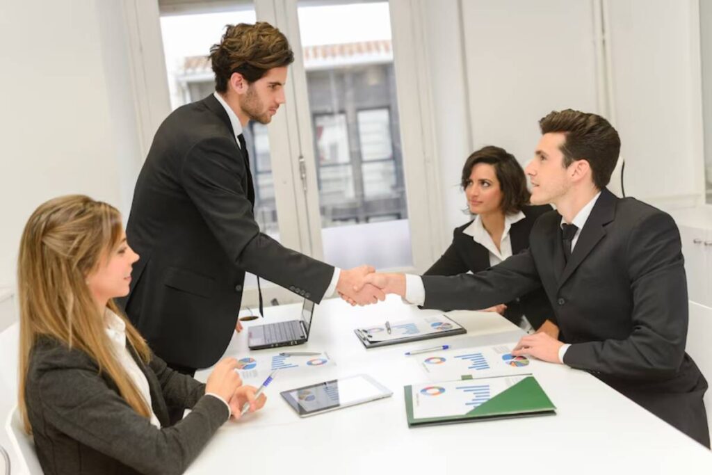 A professional meeting in a bright office; individuals shake hands while discussing documents with charts and graphs on the table.
