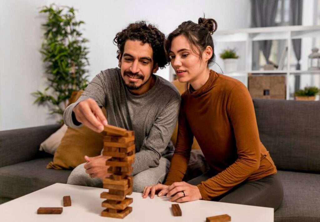 A couple is focused on a game of Jenga, carefully removing a wooden block from a tall, unstable tower in a cozy living room setting.