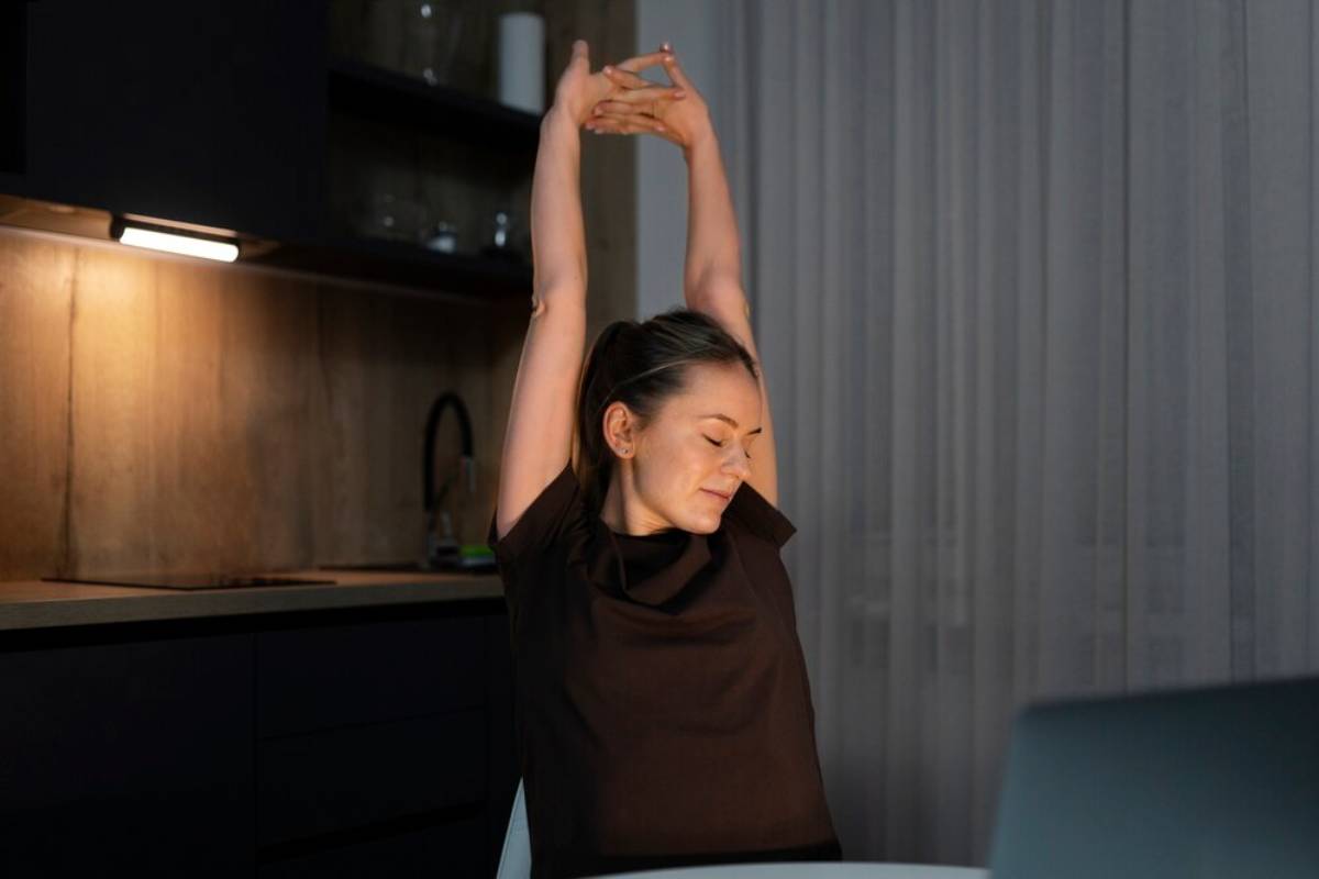 Woman stretching her arms above her head near a laptop in a dimly lit kitchen.