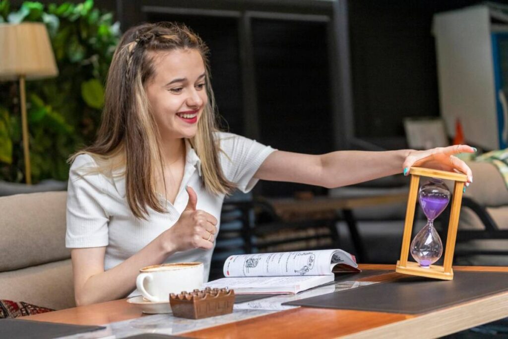 A person sits at a table with an hourglass, giving a thumbs up, a cup of coffee, and a magazine nearby in a cozy setting.