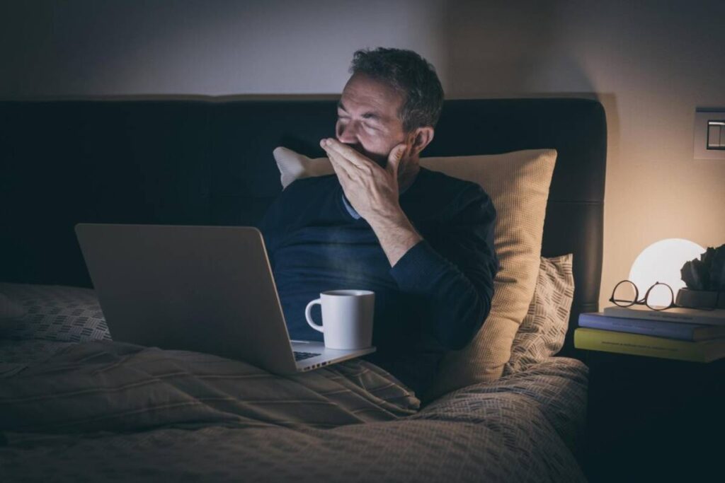 Man yawning while using a laptop in bed at night, with a coffee mug and glasses nearby.