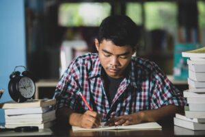Young man studying intently with rows of books and a clock beside him.