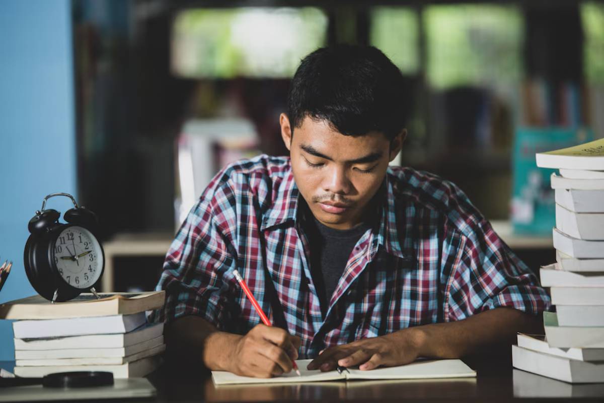 Young man studying intently with rows of books and a clock beside him.