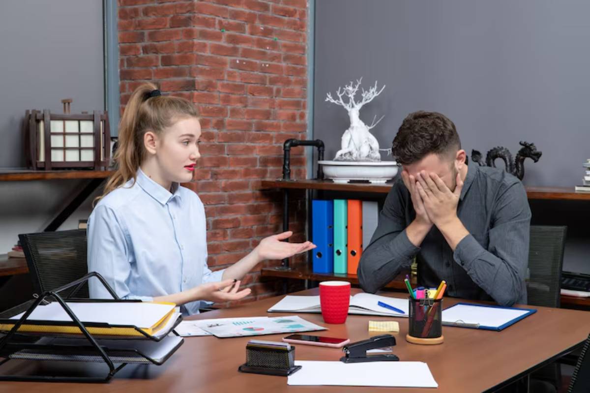 Woman talks to distressed man covering face, in a modern office with brick wall backdrop.